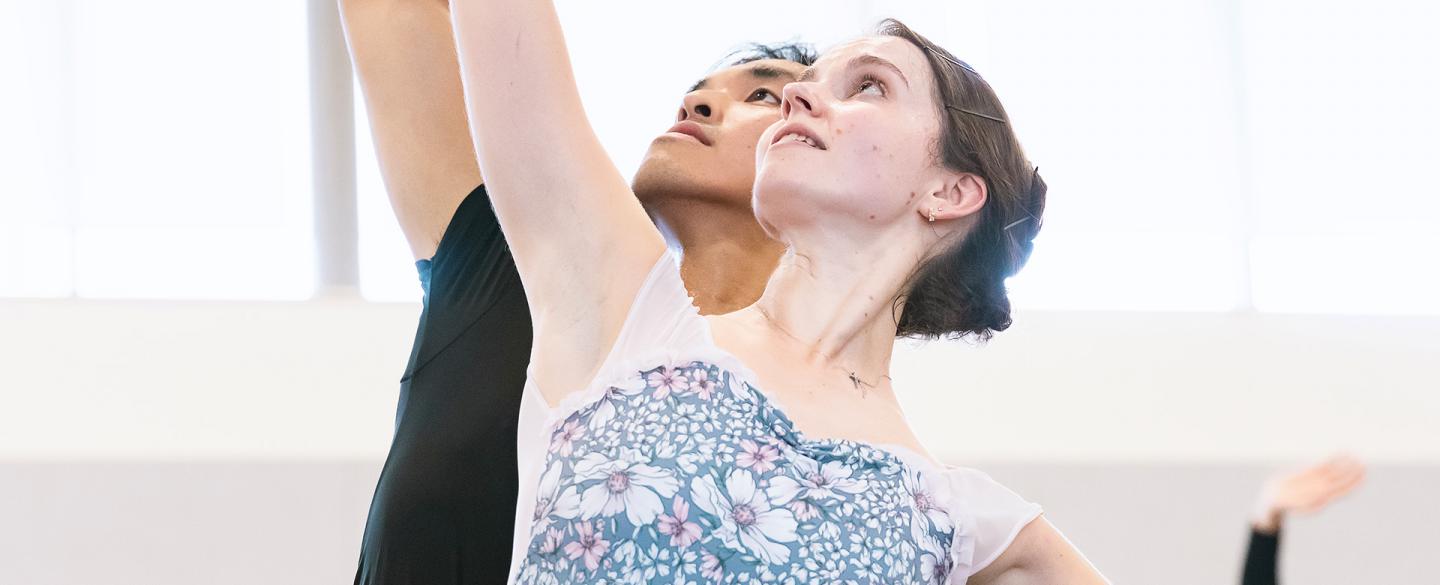 Two dancers stand with their bodies facing the camera, one behind the other, both looking up, leaning slightly back with an arm raised