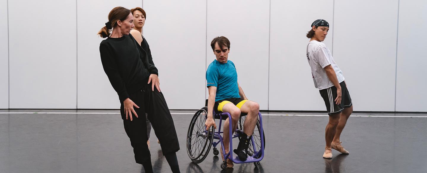 A disabled dancer in a wheelchair poses intently with two dancers and the choreographer each holding a strained pose by him