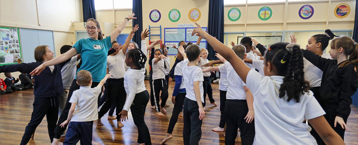 A group of children in a school hall, all barefoot, striking the same dance pose as each other - stretching one arm up and forward, the other down behind them as they step forward, their weight on the front foot