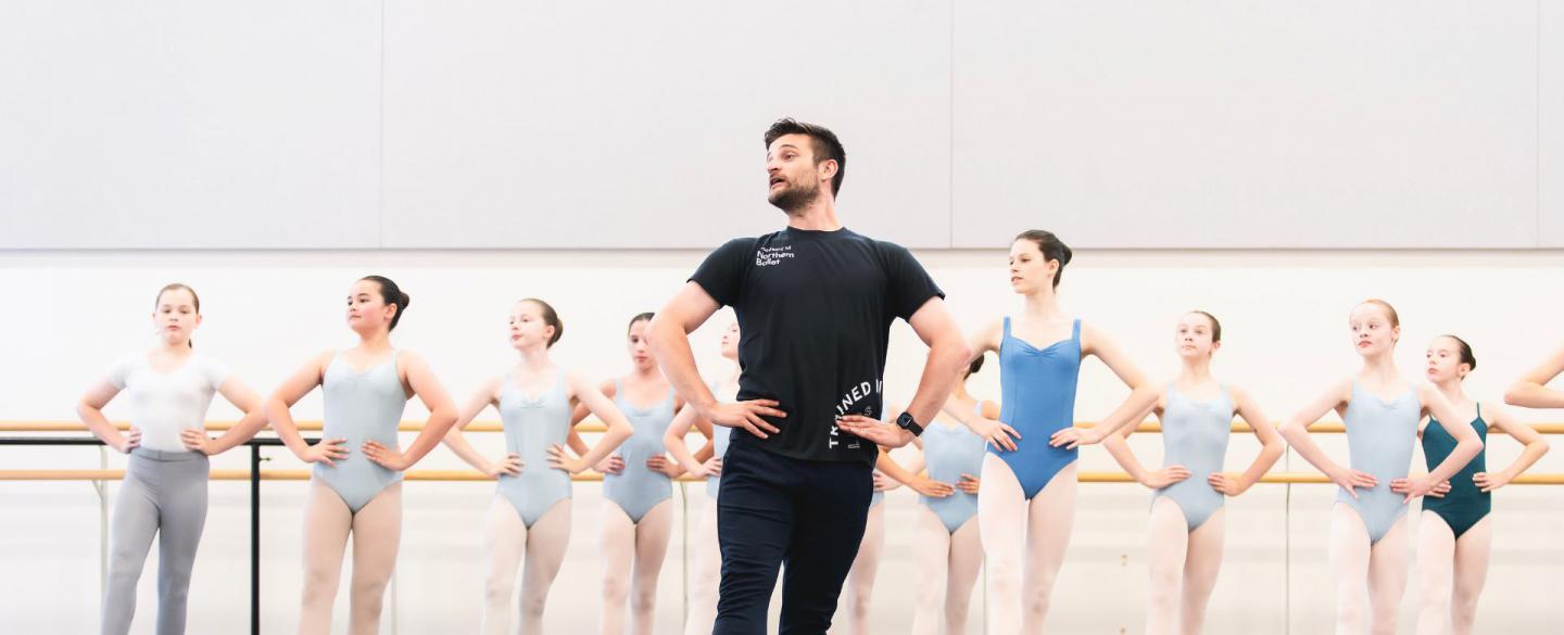 A group of students in a ballet studio stand in a line in front of a barre with their hands on their hips. They're following the lead of teacher Nicola Gervasi who stands in front of them