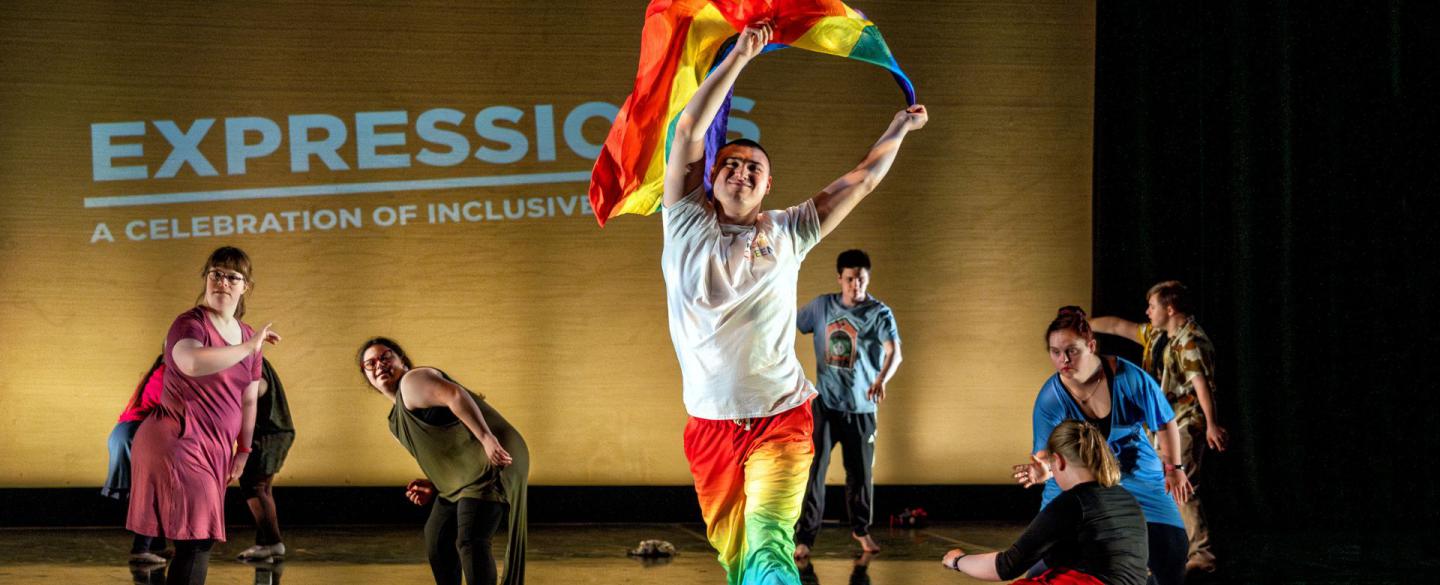 A group of performers on a stage. One strides through the centre holding a pride flag above their head.