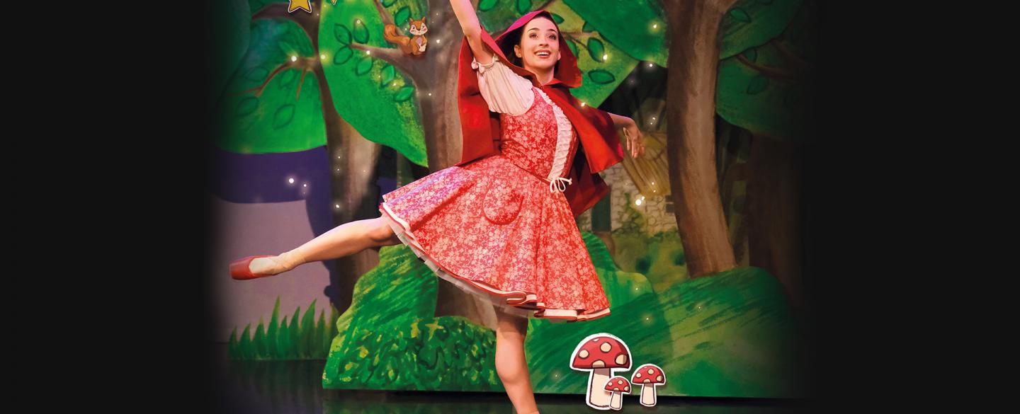 Dancer in red with a red hood and red pointe shoes stands arabesque smiling as she walks through a forest