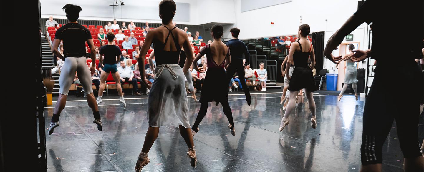 Dancers on stage in Company class, each in first jumping inches off the ground being watched by supporters