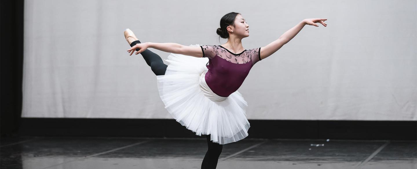 A dancer stood on one leg with the other raised and arm extended. The dancer is wearing a dark purple leotard, black leggings and a white tutu