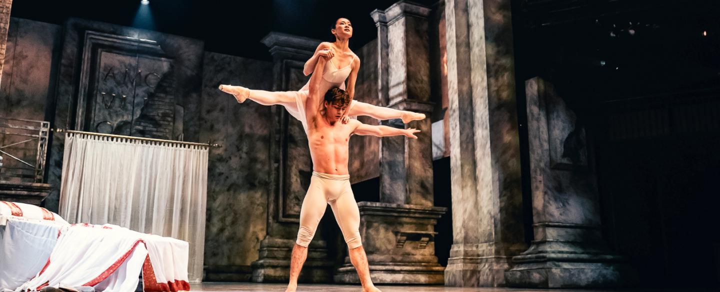 A dancer in white trousers stand in a strong pose, lifting another dancer in a white dress over his head.
