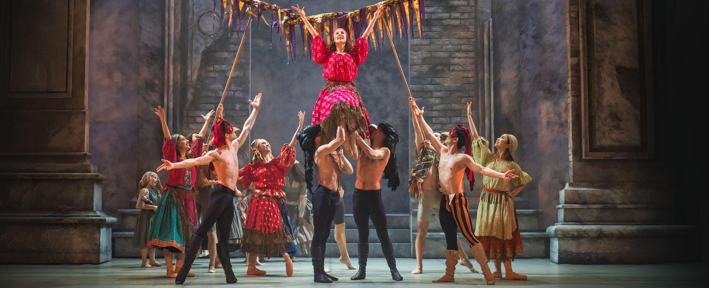 Men in masks and black leggings lift a woman in a pink patterned dress and she raises her arms in celebration