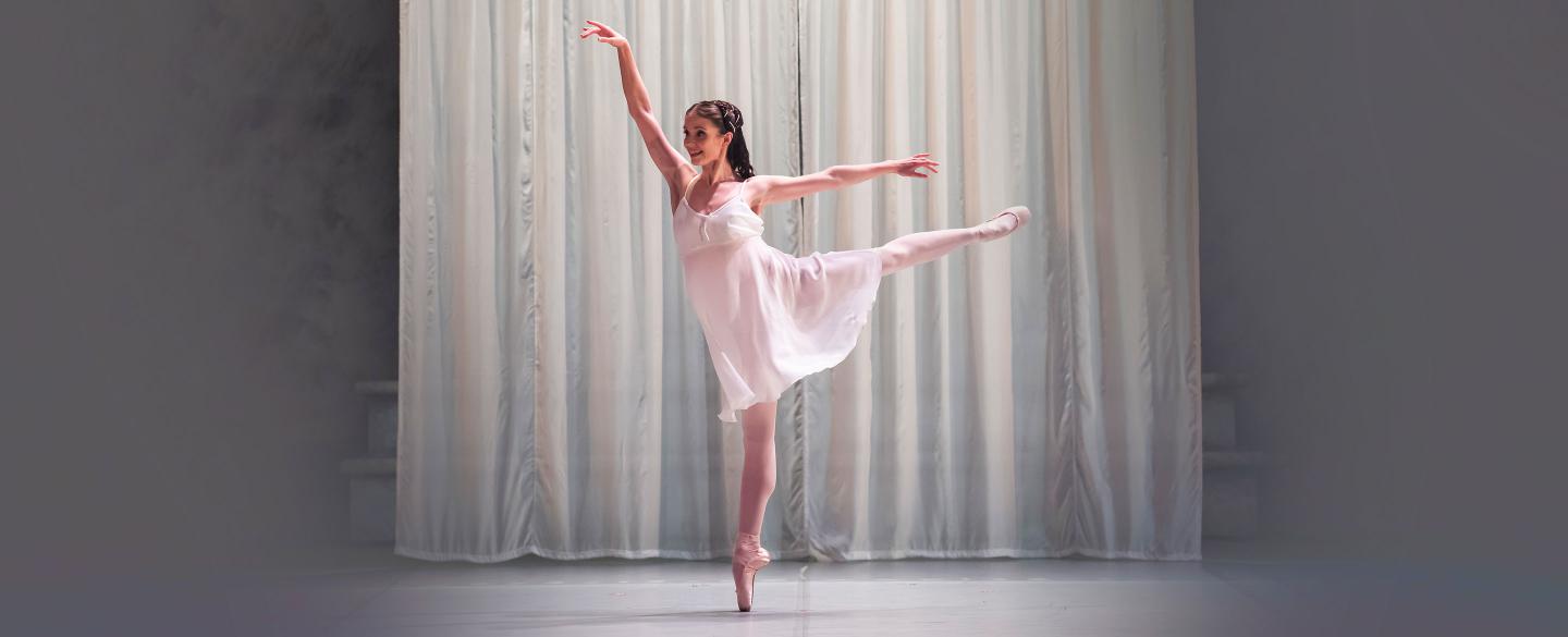 A dancer in a white dress stands on pointe with arms and legs outstretched