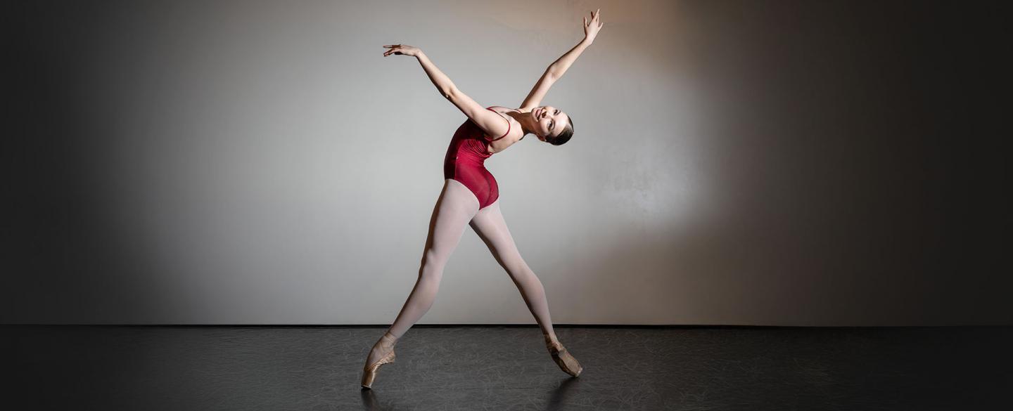Student wearing a red leotard and white tights, en pointe, her back arched back, arms raised high, smiling at the camera