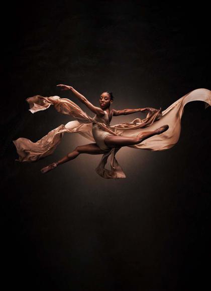 Dancer leaping across the frame while looking directly at the viewer, wearing peach and with fabrics flowing in the wind