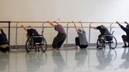 Two people in wheelchairs and three people standing, all stretching against a ballet barre