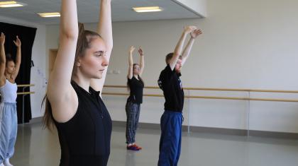 Students in a dance studio, stood still with arms raised above their heads 