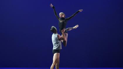 A female dancer in black leotard is lifted in the air by a male dance in black trunks and grey vest.