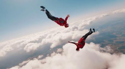 Two skydivers in red jackets and white helmets fall through the sky