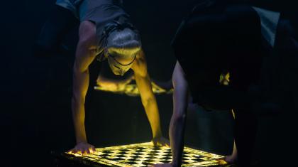 Two masked dancers look down at an illuminated chess board