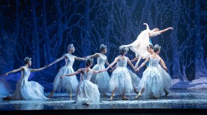 Dancers dressed in white in a whiter scene hold a young, happy woman aloft