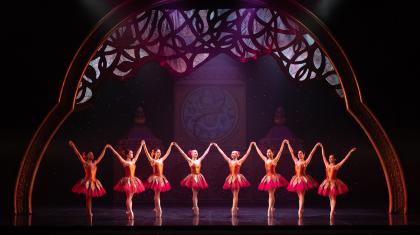 Dancers all dressed in pink and orange bodices and skirts stand in a line on their toes and arms high in the air