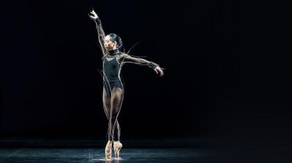 Dancer in a black leotard stands en pointe against a black background with one arm extended above her and one to her side.
