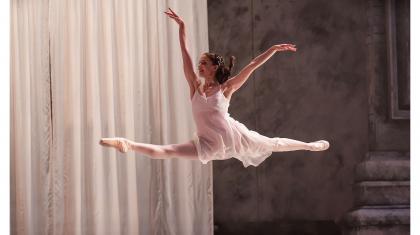 Dancer wearing white flies across the stage, leaping in a grande jete