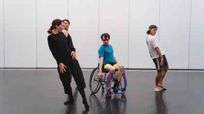 A disabled dancer in a wheelchair poses intently with two dancers and the choreographer each holding a strained pose by him