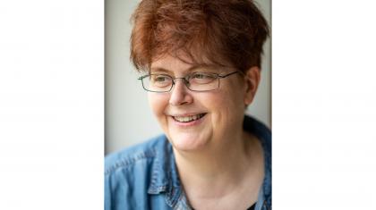 Person with dark red hair, wearing glasses, a denim shirt, smiling.