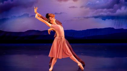 Two dancers against a cloudy background move to the right while leaning backwards and lifting their arm above their heads.