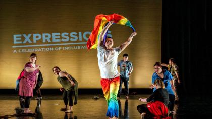 A group of performers on a stage. One strides through the centre holding a pride flag above their head.