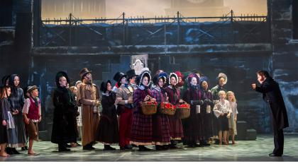 A large group of dancers in Dickensian costumes on stage singing