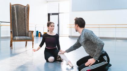 Two dancers rehearsing in a white studio, both sat on the floor. One is reaching towards the other.
