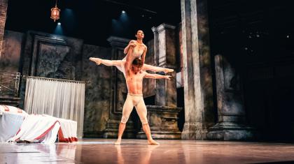 A dancer in white trousers stand in a strong pose, lifting another dancer in a white dress over his head.