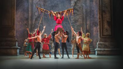 Men in masks and black leggings lift a woman in a pink patterned dress and she raises her arms in celebration