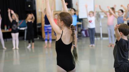 Girl in black leotard stands in a dance studio with her arms raised, in the background more children stand with their arms raised
