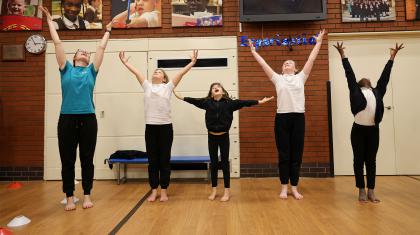 Five young people in a school hall stand on their toes, arms and heads raised joyously