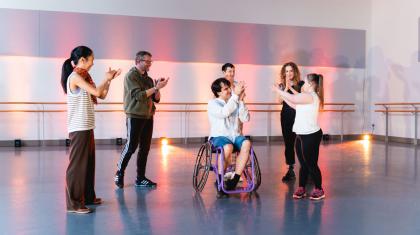 Six people stand infront of warm pink light facing each other, they are applauding.