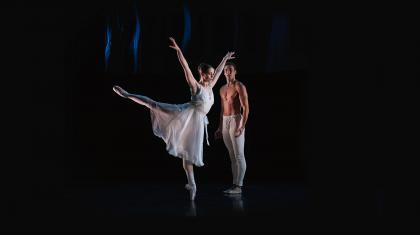 Woman in a white dress, en pointe on one foot, the other leg arabesque, both arms raise, man in white trousers stood behind her, watching.