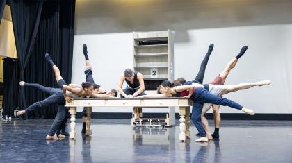 A dancer in black sits on a wooden table, leaning over whilst writing with a feather quill. Multiple dancers surround the table, each with one leg elevated behind them, reaching towards the quill.
