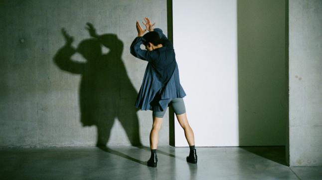 A man in a blue oversized blouse and black boots forms a shadow puppet with his arms against a concrete wall
