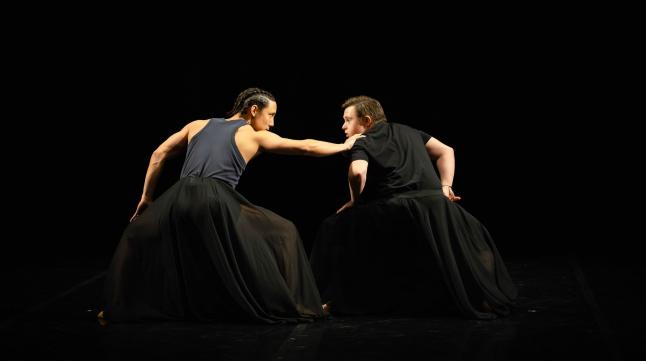 Two dancers stand hands on their bent knees. They look at each other as one grabs the other's shoulder.